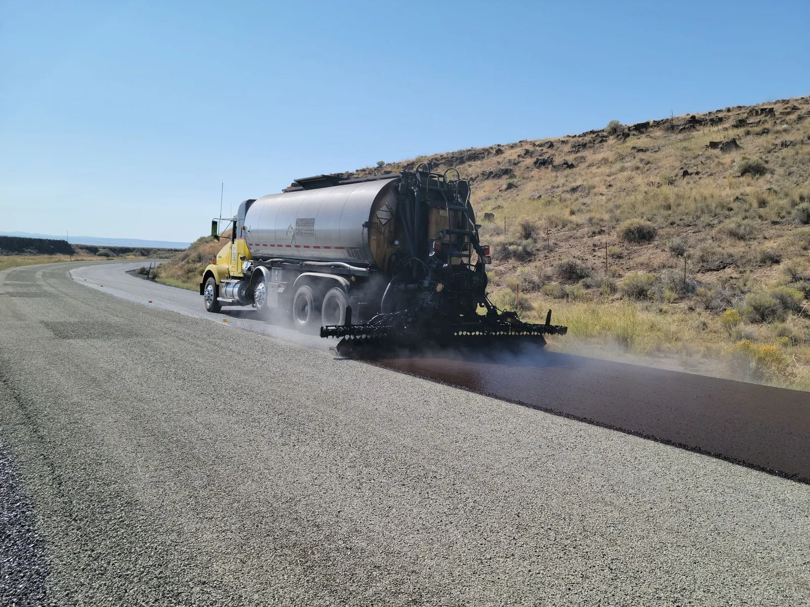 A road tanker applying asphalt on a rural road in Oregon under clear skies.