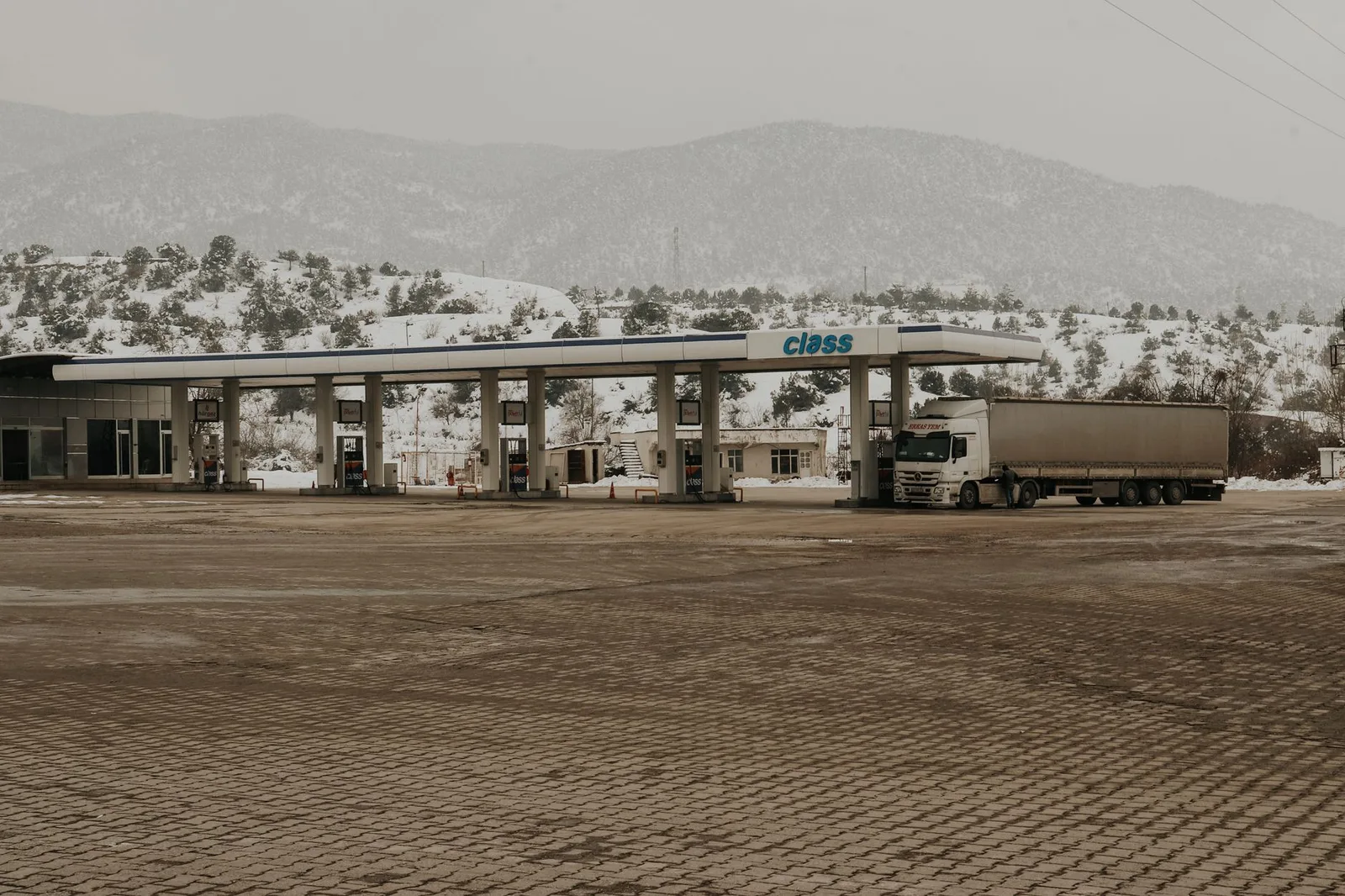 A snowy gas station with a truck and mountains in the backdrop, offering a serene winter scene.